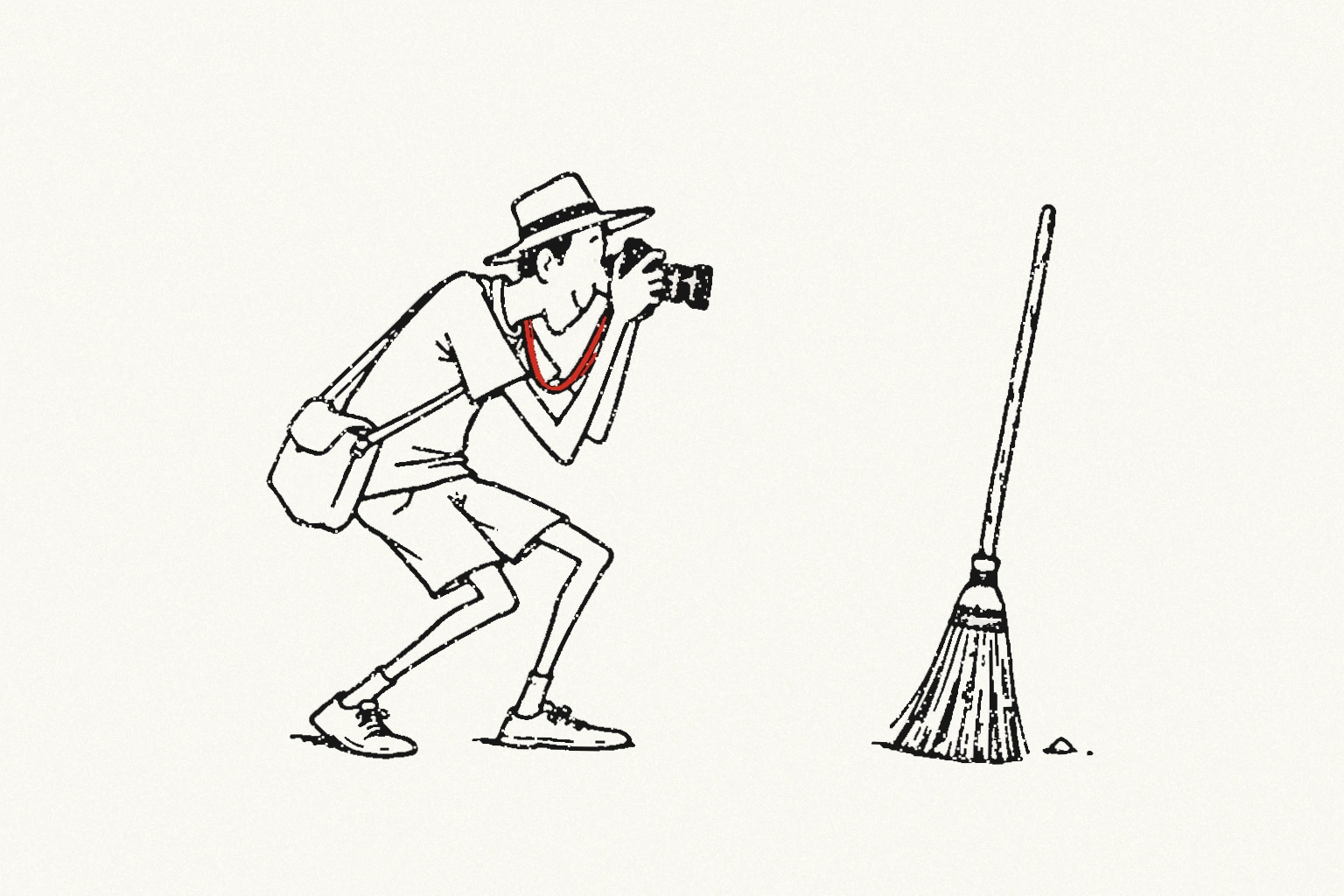 A tourist in a brimmed hat crouches with his camera in front of an ordinary broom and a tiny dust pile; his camera strap is red.